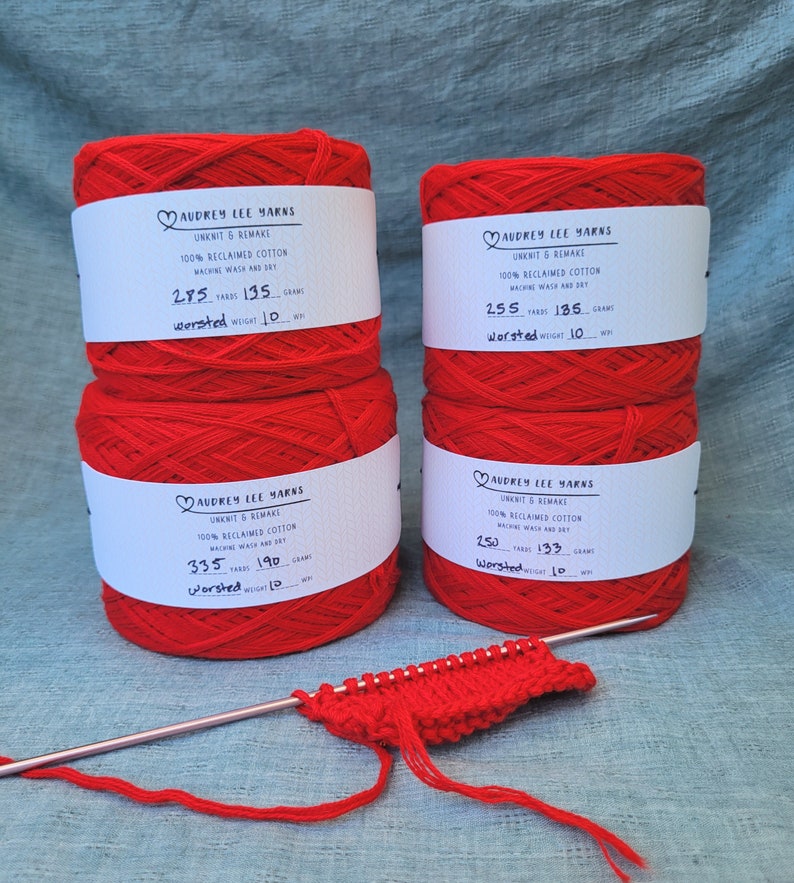 May include: Four spools of red yarn labeled "Audrey Lee Yarns" with details about the yarn, including the yarn weight, yardage, and grams. A knitting needle and a small swatch of red knit fabric are also pictured.
