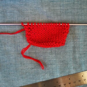 May include: Red knitted fabric on a knitting needle. The fabric is on a blue surface with a ruler in the bottom right corner.