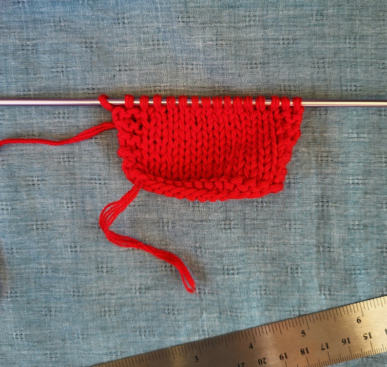 May include: Red knitted fabric on a knitting needle. The fabric is on a blue surface with a ruler in the bottom right corner.