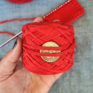 May include: A ball of red yarn with a US quarter dollar coin on top. The yarn is wrapped around the coin. The yarn is in a person's hand.