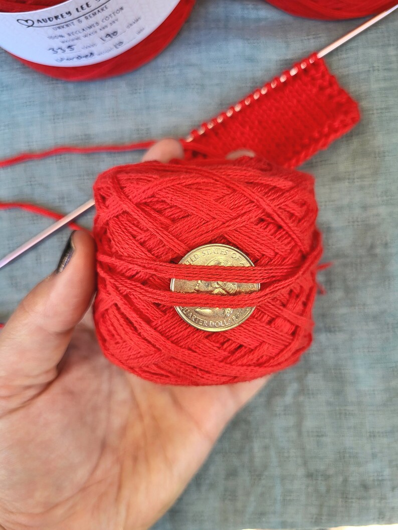 May include: A ball of red yarn with a US quarter dollar coin on top. The yarn is wrapped around the coin. The yarn is in a person's hand.