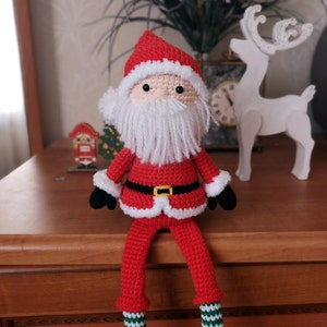 May include: A crocheted Santa Claus doll featuring a red suit with white trim, a black belt with a yellow buckle, black mittens, and a long white yarn beard. The doll has a beige face with black bead eyes and wears a red hat with white trim.