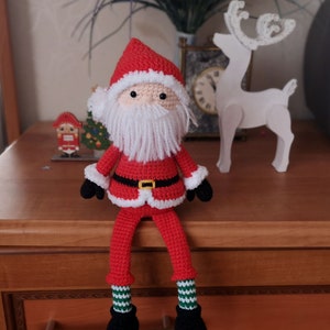May include: A handmade crochet Santa Claus doll featuring a red hat and coat with white fuzzy trim, a long white yarn beard, black gloves, and black boots. The doll has long legs with green and white striped socks and a black belt with a yellow buckle.