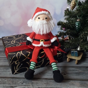 May include: A crocheted Santa Claus doll featuring a red suit with white trim, a black belt with a yellow buckle, black gloves, and black boots. The doll has a white yarn beard, a red hat, and green and white striped legs, sitting upright.
