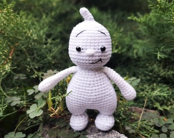 May include: White crocheted ghost amigurumi toy with black eyes and a smiling mouth. The toy is sitting on a rock with green foliage in the background.