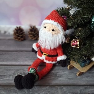 May include: A crocheted Santa Claus doll featuring a red hat and suit with white trim, a long white yarn beard, a black belt with a yellow buckle, black mittens, and black boots with green and white patterned socks, sitting upright.
