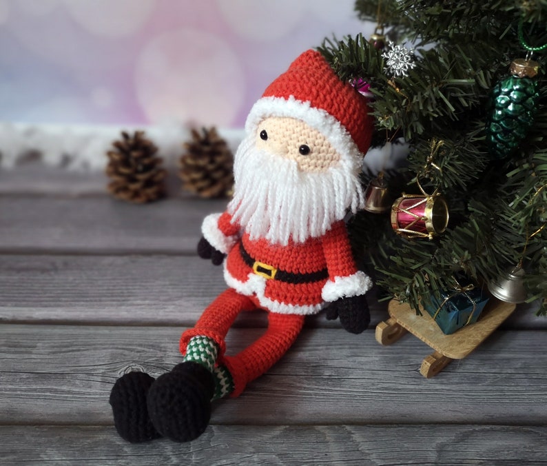May include: A crocheted Santa Claus doll featuring a red hat and suit with white trim, a long white yarn beard, a black belt with a yellow buckle, black mittens, and black boots with green and white patterned socks, sitting upright.