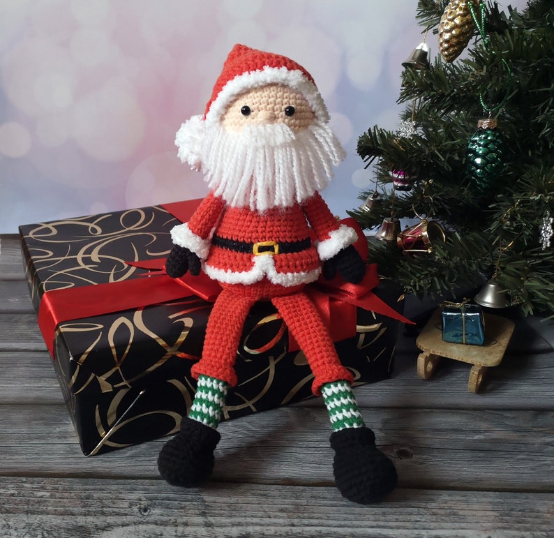 May include: A crocheted Santa Claus doll featuring a red suit with white trim, a black belt with a yellow buckle, black gloves, and black boots. The doll has a white yarn beard, a red hat, and green and white striped legs, sitting upright.