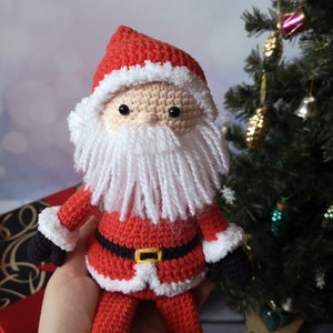 May include: A crocheted Santa Claus doll featuring a red hat with white trim, a beige face with black bead eyes, and a long white yarn beard. The doll wears a red suit with a black belt, a yellow buckle, white cuffs, and black mittens on its hands.