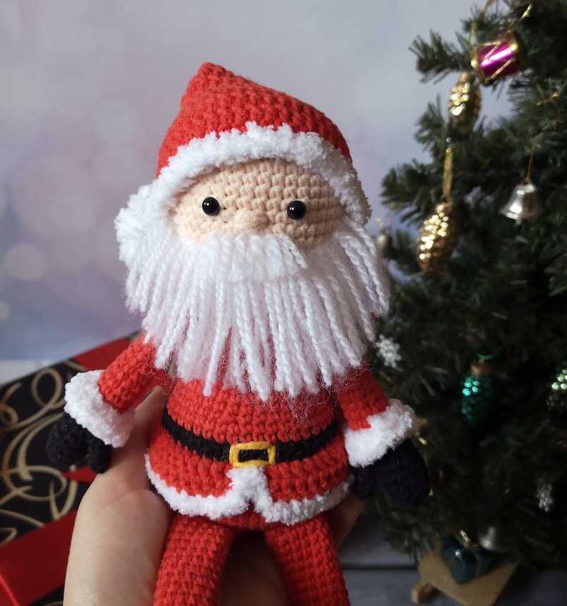 May include: A crocheted Santa Claus doll featuring a red hat with white trim, a beige face with black bead eyes, and a long white yarn beard. The doll wears a red suit with a black belt, a yellow buckle, white cuffs, and black mittens on its hands.