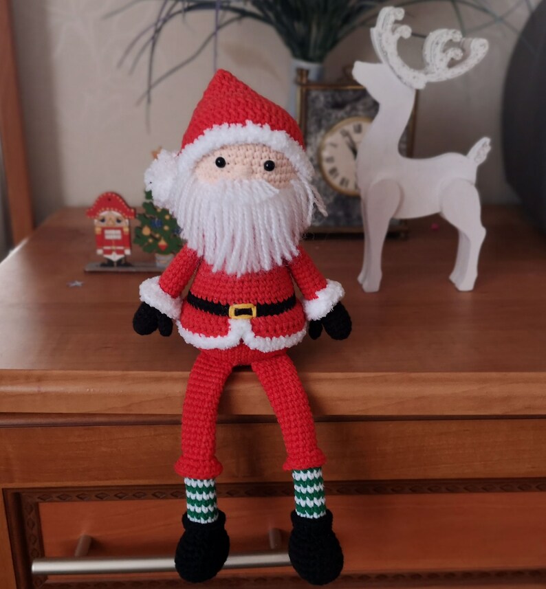 May include: A handmade crochet Santa Claus doll featuring a red hat and coat with white fuzzy trim, a long white yarn beard, black gloves, and black boots. The doll has long legs with green and white striped socks and a black belt with a yellow buckle.