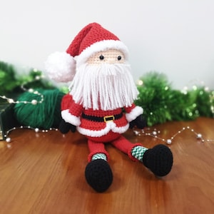 May include: A crocheted Santa Claus doll featuring a red hat with a white pom-pom, a long white yarn beard, a red jacket with a black belt and gold buckle, and black boots. The doll has green and white chequered socks and small black eyes.