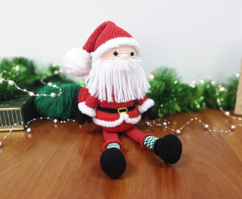 May include: A crocheted Santa Claus doll featuring a red hat with a white pom-pom, a long white yarn beard, a red jacket with a black belt and gold buckle, and black boots. The doll has green and white chequered socks and small black eyes.