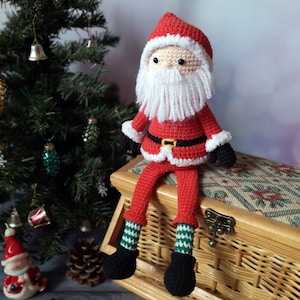 May include: A crocheted Santa Claus doll featuring a red hat and coat with white trim, a black belt with a yellow buckle, black mittens, and black boots. The doll has a white yarn beard, a peach-coloured face, and green and white striped socks.