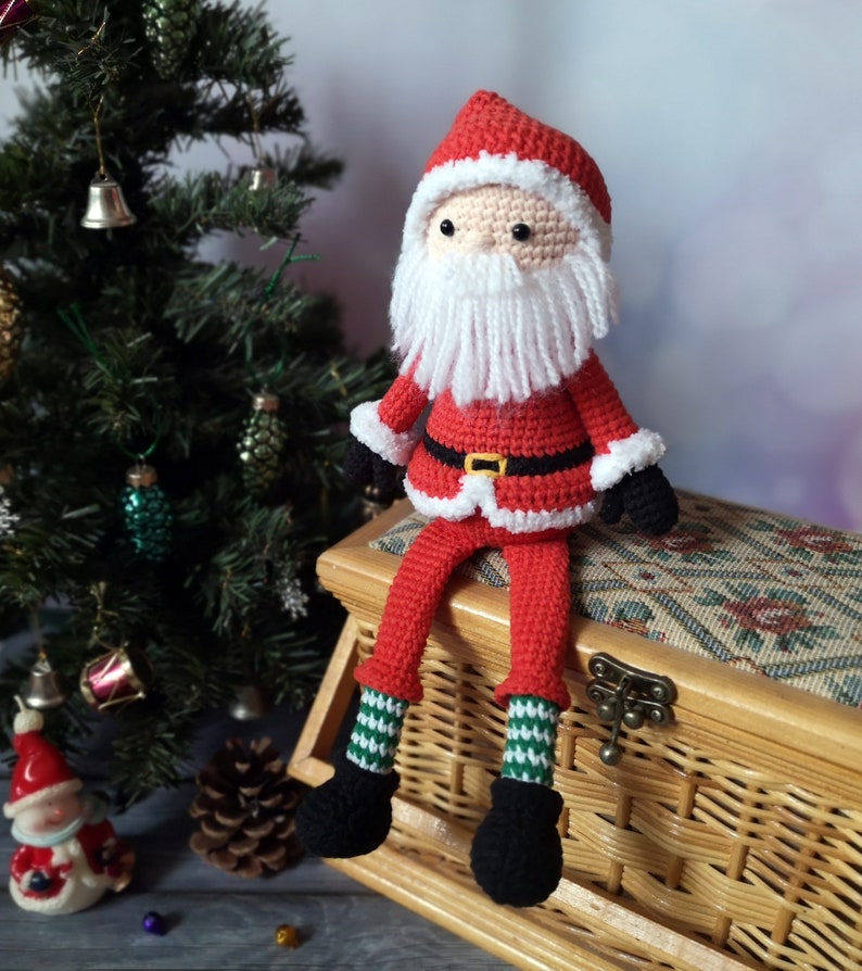 May include: A crocheted Santa Claus doll featuring a red hat and coat with white trim, a black belt with a yellow buckle, black mittens, and black boots. The doll has a white yarn beard, a peach-coloured face, and green and white striped socks.