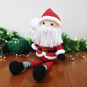 May include: A crocheted Santa Claus doll featuring a red hat with white trim, a long white yarn beard, a red jacket with a black belt and gold buckle, green and white patterned legs, and black boots. The doll has small black bead eyes.
