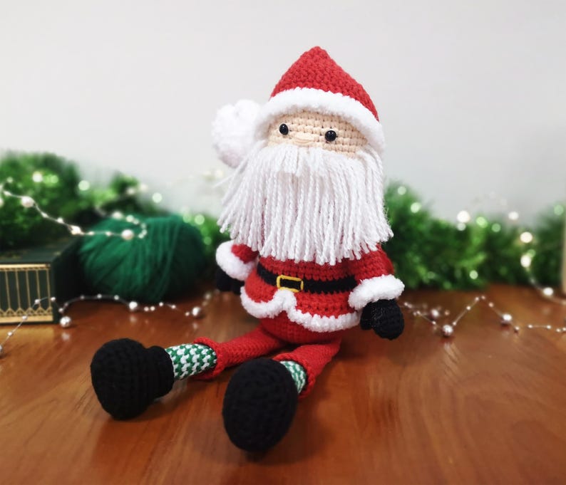 May include: A crocheted Santa Claus doll featuring a red hat with white trim, a long white yarn beard, a red jacket with a black belt and gold buckle, green and white patterned legs, and black boots. The doll has small black bead eyes.