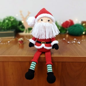 May include: A crocheted Santa Claus doll featuring a red hat with a white pom-pom, a long white yarn beard, a red jacket with white trim and a black belt, and long legs with green and white striped stockings ending in black crocheted boots.