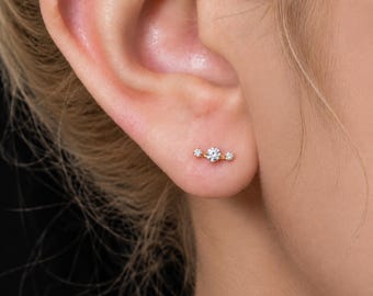 May include: A gold stud earring featuring a central round clear stone flanked by two smaller clear stones on each side, arranged in a horizontal line. The earring is secured in the earlobe, showcasing a delicate metal setting and clear gems.