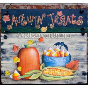 Op de afbeelding: Een houten bord met de tekst "Autumn Treats" geschilderd in oranje en bruin. Het bord is versierd met herfstbladeren en een pompoen. Onder het bord is een geschilderd beeld van een pompoen, maïs en een kom met candy corn met een vleermuis erop. De kom is blauw en er staat "Candy Corn" op.