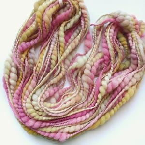 May include: A skein of hand-spun art yarn featuring a textured blend of pink, cream, and pale yellow fibres. The yarn consists of multiple strands with varying thicknesses, including thick coiled sections, thin plied threads, and soft roving.