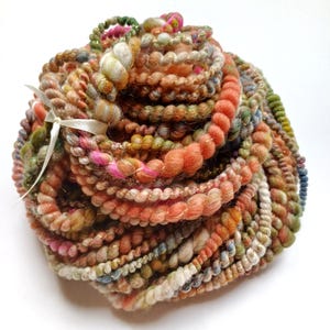 May include: A large, round skein of textured art yarn featuring a variegated color palette of peach, orange, pink, green, and cream. The yarn has a thick, coiled structure with metallic gold thread accents and is tied with a thin white ribbon.