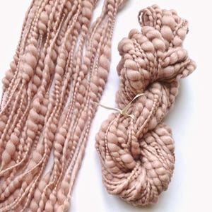 May include: Close-up of two types of blush-colored yarn. One is a skein of chunky, textured yarn tied with a cream-colored ribbon. The other is a series of long, thick strands of yarn with a braided appearance.