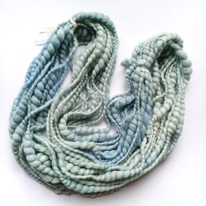 May include: A skein of thick, textured yarn featuring a gradient of soft blue and pale mint green hues. The yarn has a distinct bobble or knot-like structure throughout its length, creating a varied, bumpy surface texture across the entire bundle.