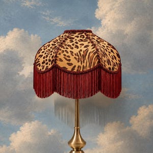 May include: A table lamp featuring a brass-toned metal base and a scalloped lampshade with a leopard print pattern. The lampshade is trimmed with deep red fringe and matching braided cord along the top and bottom edges.