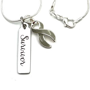 Può includere: Collana con catena d'argento con un ciondolo d'argento che dice "Survivor" e un ciondolo a nastro grigio.