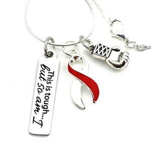 May include: Silver necklace with three charms: a red and white ribbon, a silver boxing glove, and a silver tag with the text "This is tough... but so am I."