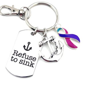 Puede incluir: Llavero plateado con un dije de ancla plateado, un dije de cinta azul, rosa y turquesa, y una etiqueta plateada que dice "Refuse to sink".
