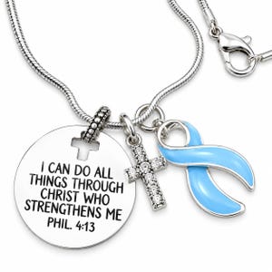 May include: A silver-toned snake chain necklace featuring three charms: a round metal disc engraved with the text I CAN DO ALL THINGS THROUGH CHRIST WHO STRENGTHENS ME PHIL. 4:13, a small cross pendant encrusted with clear stones, and a light blue ribbon.