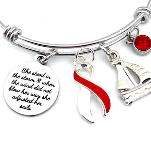 Può includere: Un braccialetto con ciondoli in argento con un ciondolo a nastro rosso e bianco, un ciondolo a barca a vela e un ciondolo rotondo con la scritta "She stood in the storm & when the wind did not blow her way she adjusted her sails".