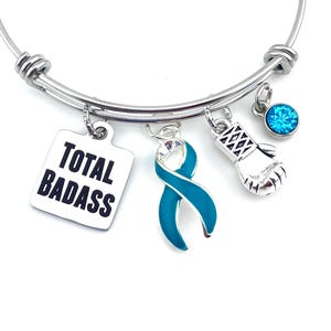 Op de afbeelding: Een uitbreidbare armband van zilverkleurig draad met vier hangende bedels: een vierkante zilveren hanger met de zwarte tekst TOTAL BADASS, een blauwgroen bewustzijnslintje, een zilveren bokshandschoen en een kleine ronde felblauwe kristalbedel.