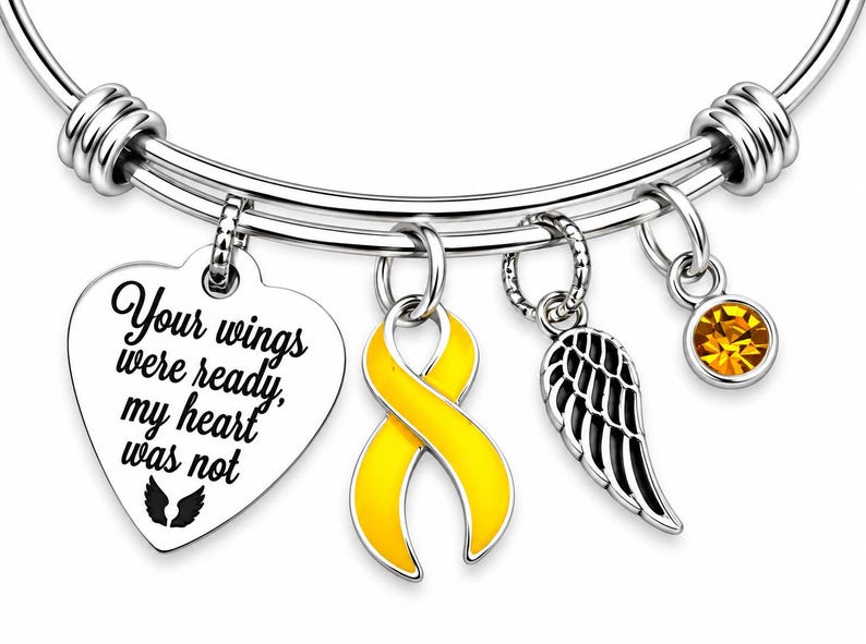 May include: A silver-tone bangle bracelet featuring four charms: a heart-shaped pendant engraved with the text "Your wings were ready, my heart was not" and a small wing icon, a yellow awareness ribbon, a detailed angel wing, and a yellow crystal.