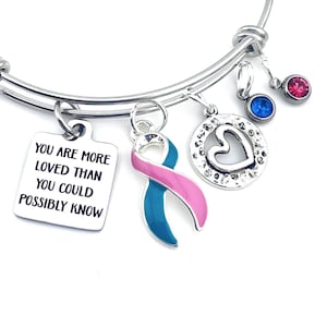 May include: A silver charm bracelet with a teal and pink ribbon charm, a heart charm, and a charm with the text "You are more loved than you could possibly know".