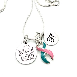 May include: Silver necklace with a teal and pink ribbon charm, a round charm with the text "She Believed She Could So She Did", and a small charm with the letter "J".