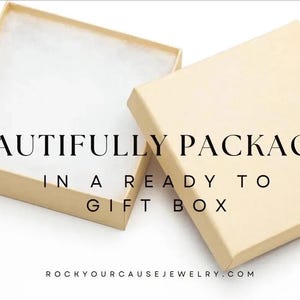 May include: A square, light beige cardboard gift box with a removable lid placed beside the open base, which contains white cotton padding. The text "BEAUTIFULLY PACKAGED IN A READY TO GIFT BOX" and "ROCKYOURCAUSEJEWELRY.COM" is printed on the image.