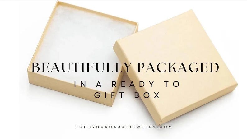 May include: A square, light beige cardboard gift box with a removable lid placed beside the open base, which contains white cotton padding. The text "BEAUTIFULLY PACKAGED IN A READY TO GIFT BOX" and "ROCKYOURCAUSEJEWELRY.COM" is printed on the image.