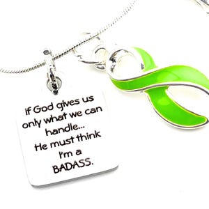 May include: A silver-toned necklace featuring a square pendant engraved with the text 'If God gives us only what we can handle... He must think I'm a BADASS.' and a separate lime green awareness ribbon charm attached to the thin silver chain.