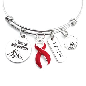 May include: Silver-toned bangle bracelet with multiple charms. Charms include a red ribbon, a tag with the word "FAITH", a round charm with a mountain scene, and a round charm with the text "Faith Can Move Mountains".