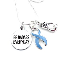 May include: Silver necklace with three charms: a round charm with the text "BE BADASS EVERYDAY", a blue ribbon charm, and a silver boxing glove charm.