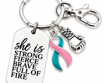 Könnte beinhalten: Ein silberfarbener Metallschlüsselanhänger mit einem rechteckigen Anhänger, der mit dem Text 'she is STRONG FIERCE BRAVE FULL OF FIRE' graviert ist. Daran befestigt sind zwei Anhänger: eine rosa-türkisfarbene Schleife und ein kleiner Boxhandschuh.