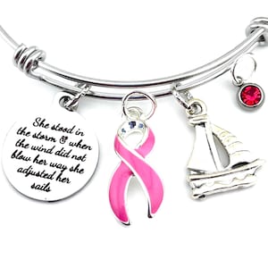May include: A silver charm bracelet with a pink ribbon charm, a sailboat charm, a red crystal charm, and a charm with the text "She stood in the storm & when the wind did not blow her way she adjusted her sails".
