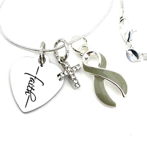Op de afbeelding: Zilveren ketting met een hartvormige hanger met de tekst "Faith", een klein kruisje met strass steentjes en een groene lint hanger.