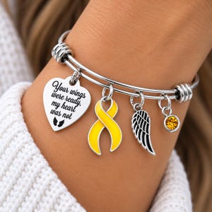 May include: A silver-toned expandable bangle bracelet featuring four charms: a heart-shaped pendant engraved with the text "Your wings were ready, my heart was not" and two small wings, a yellow awareness ribbon charm, a black wing charm, and a yellow crystal.