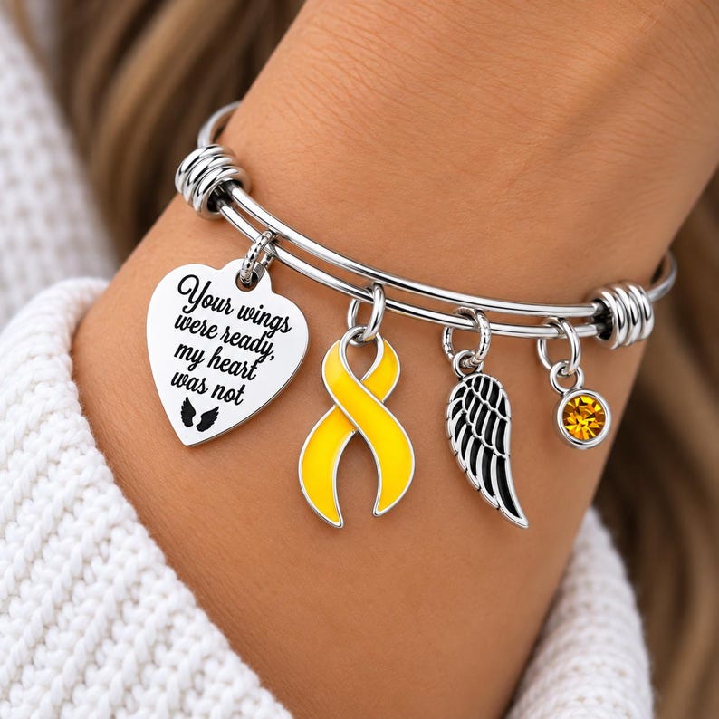 May include: A silver-toned expandable bangle bracelet featuring four charms: a heart-shaped pendant engraved with the text "Your wings were ready, my heart was not" and two small wings, a yellow awareness ribbon charm, a black wing charm, and a yellow crystal.
