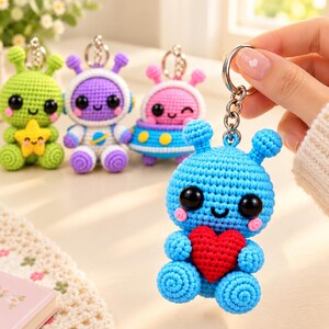 May include: A blue crochet keychain featuring a small creature with large black eyes, pink cheeks, and two antennae, holding a red heart. In the background, three additional crochet keychains are visible: a green one with a yellow star, a purple astronaut, and a pink creature in a blue saucer.