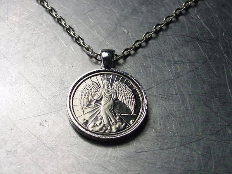 May include: Silver necklace with a round pendant featuring an angel with wings. The angel is depicted in a silver relief design.